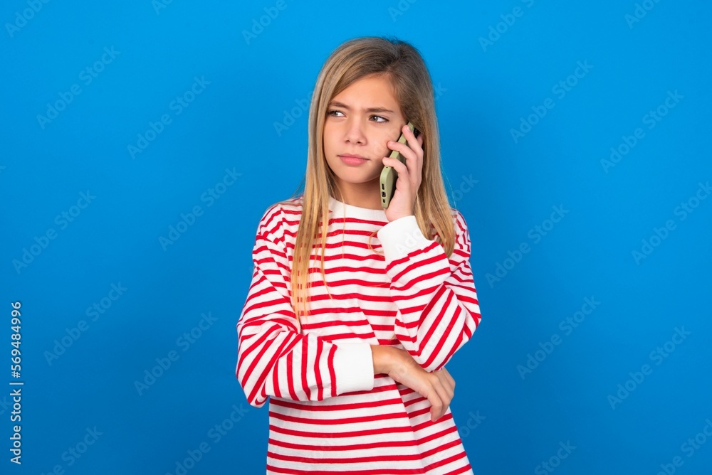 Sad beautiful caucasian teen girl wearing striped T-shirt over blue wall talking on smartphone. Communication concept. Sad beautiful caucasian teen girl wearing striped T-shirt over blue wall talking on smartphone. Communication concept.