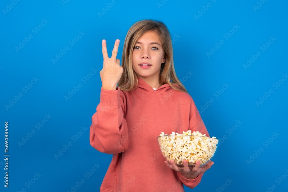 beautiful caucasian teen girl wearing pink sweater over blue wall eating popcorn smiling and looking friendly, showing number two or second with hand forward, counting down beautiful caucasian teen girl wearing pink sweater over blue wall eating popcorn smiling and looking friendly, showing number two or second with hand forward, counting down