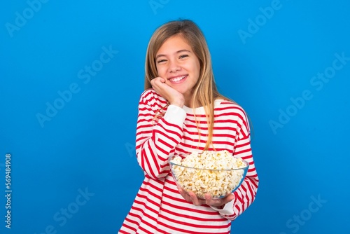 Positive beautiful caucasian teen girl wearing striped T-shirt over blue wall eating popcorn smiles happily, glad to receive pleasant news from interlocutor.