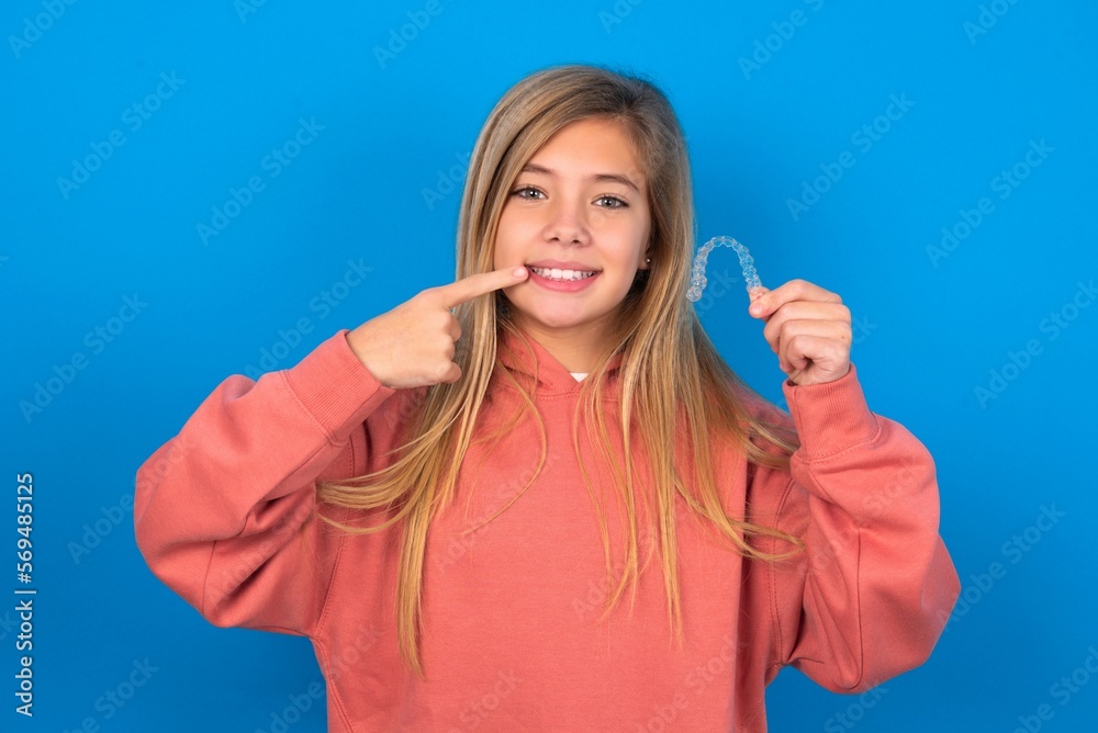 Fototapeta premium beautiful caucasian teen girl wearing pink sweater over blue wall holding an invisible aligner and pointing perfect straight teeth. Dental healthcare and confidence concept.