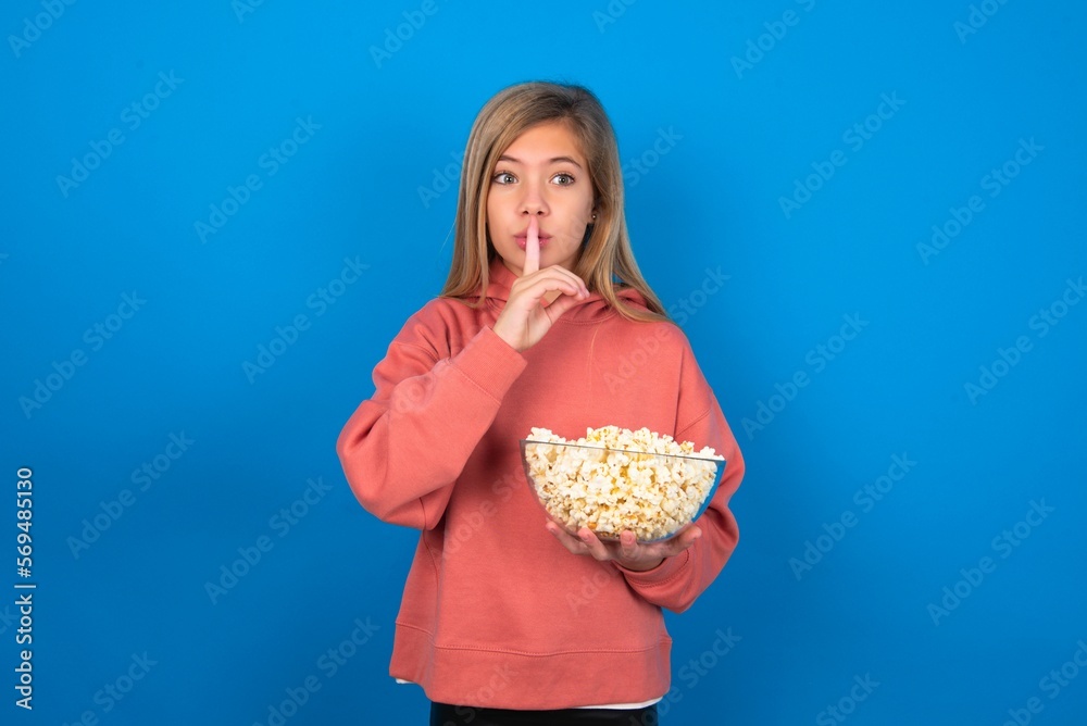 Surprised beautiful caucasian teen girl wearing pink sweater over blue wall eating popcorn makes silence gesture, keeps finger over lips and looks mysterious at camera