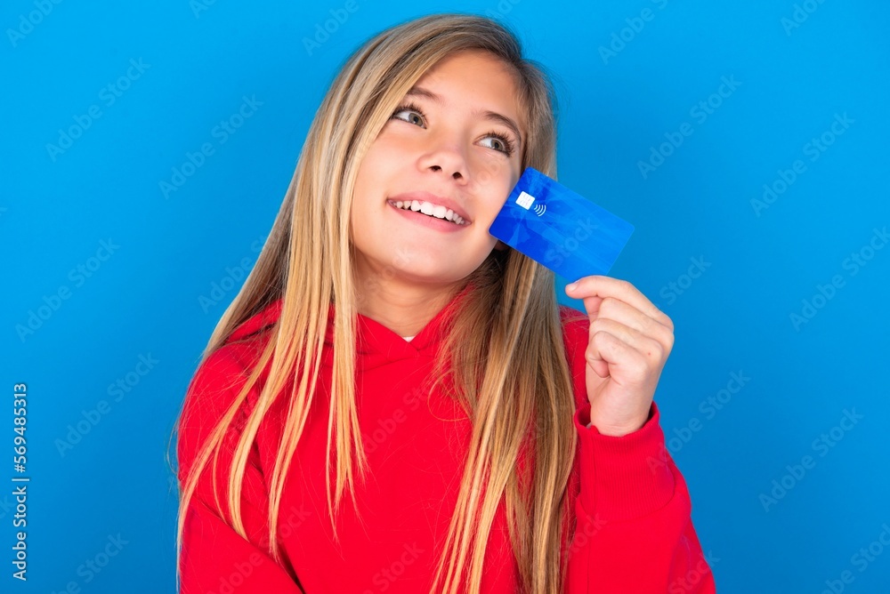 Lovely beautiful caucasian teen girl wearing red sweater over blue wall showing credit card and looking away at copy space. Pensive Lovely beautiful caucasian teen girl wearing red sweater over blue wall showing credit card and looking away at copy space. Pensive