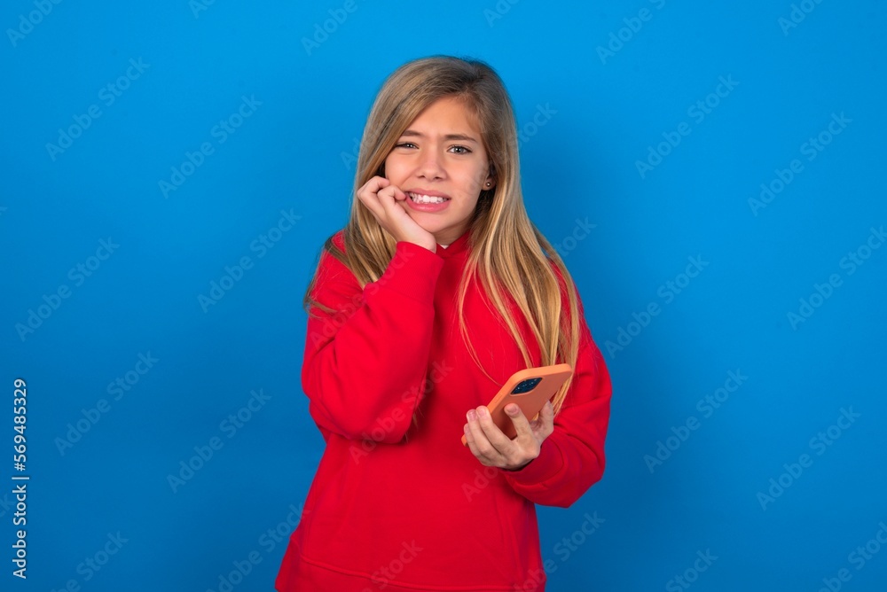 Afraid funny beautiful caucasian teen girl wearing red sweater over blue wall holding telephone and bitting nails Afraid funny beautiful caucasian teen girl wearing red sweater over blue wall holding telephone and bitting nails