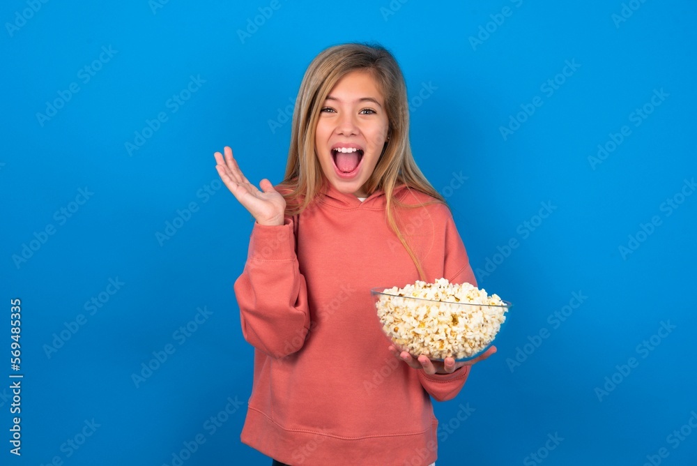 Surprised happy Smiling beautiful caucasian teen girl wearing pink sweater over blue wall, eating popcorn, keeps hands near head, jaw dropped. Surprised happy Smiling beautiful caucasian teen girl wearing pink sweater over blue wall, eating popcorn, keeps hands near head, jaw dropped.