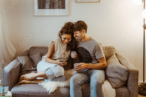 Happy couple having coffee while sharing smart phone sitting on sofa at home