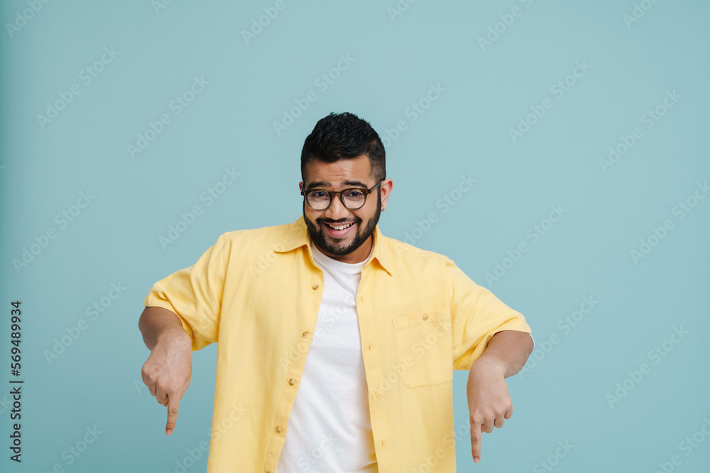 Cheerful indian man pointing fingers down at copy space isolated over blue wall Cheerful indian man pointing fingers down at copy space isolated over blue wall