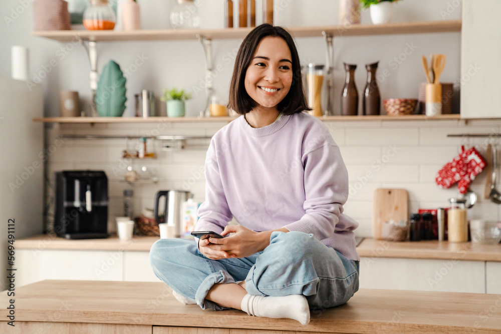 Cheerful asian woman using mobile phone while sitting on table in kitchen Cheerful asian woman using mobile phone while sitting on table in kitchen