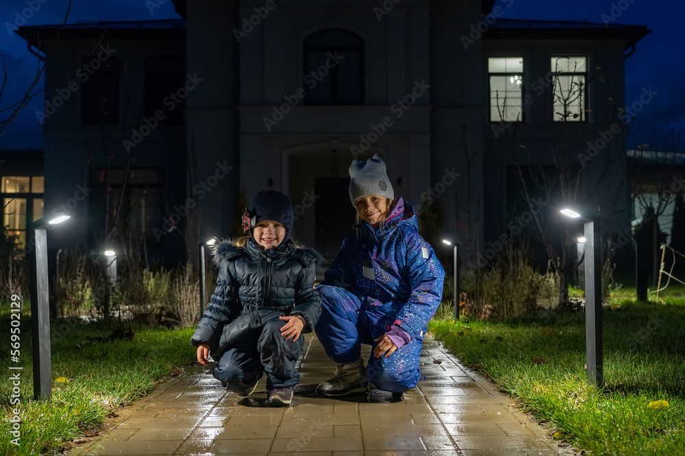 Obraz premium Two happy sisters pose on path in front of house illuminated by row of modern street lamps on rainy evening backyard