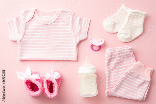 Baby shower concept. Top view photo of pink infant clothes shirt pants tiny socks pacifier milk bottle and knitted booties on isolated pastel pink background