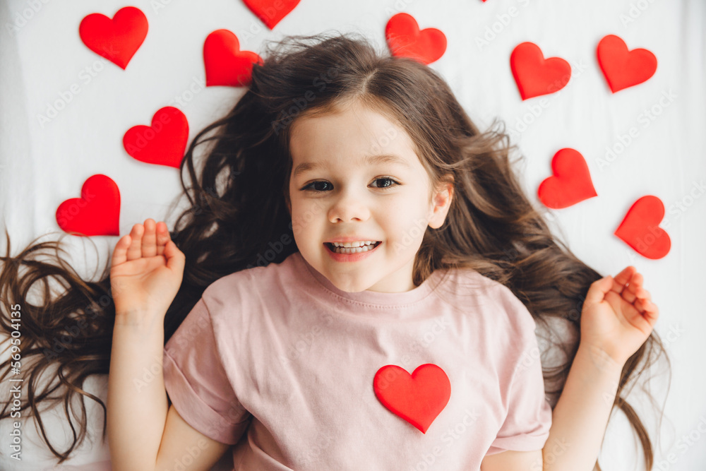 Valentine's Day, a little girl lying at home on a white bed among hearts, smiling and laughing with happiness, congratulating on the holiday Valentine's Day, a little girl lying at home on a white bed among hearts, smiling and laughing with happiness, congratulating on the holiday