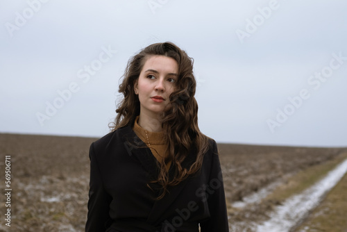 A beautiful girl is standing in a field.Natural beauty.Curly brunette.Emotions of calmness and reflection.Woman dreams.Lady in black.Portrait of a young woman.Retro and vintage.Deep beautiful look.