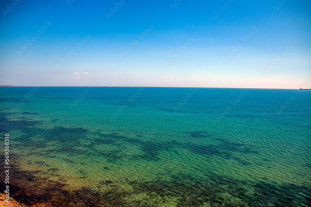 Turquoise transparent sea with algae at the bottom taken from a cliff with ships on the horizon and blue sky