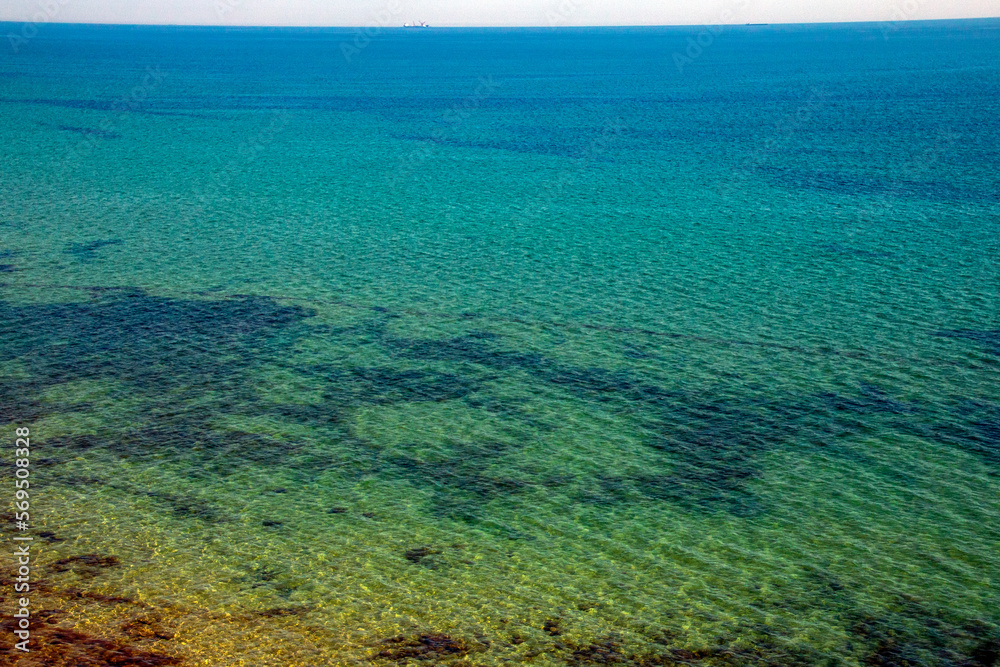 Turquoise transparent sea with algae at the bottom taken from a cliff with ships on the horizon