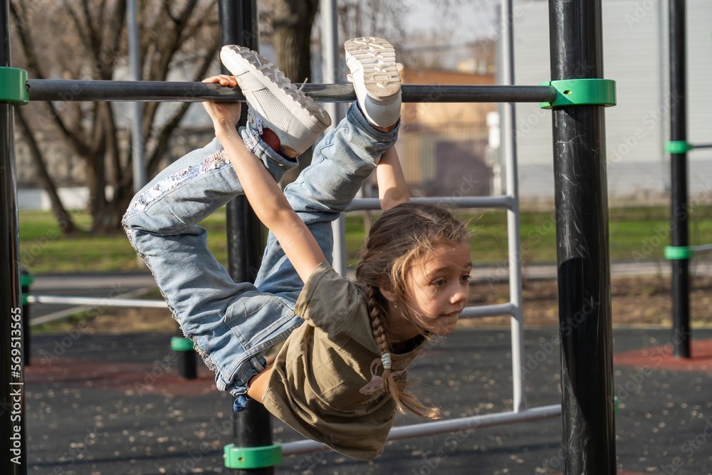 Obraz premium Happy girl in after school hanging upside down on gym equipment. Cheerful girl athlete on gymnastic stick on sports ground.