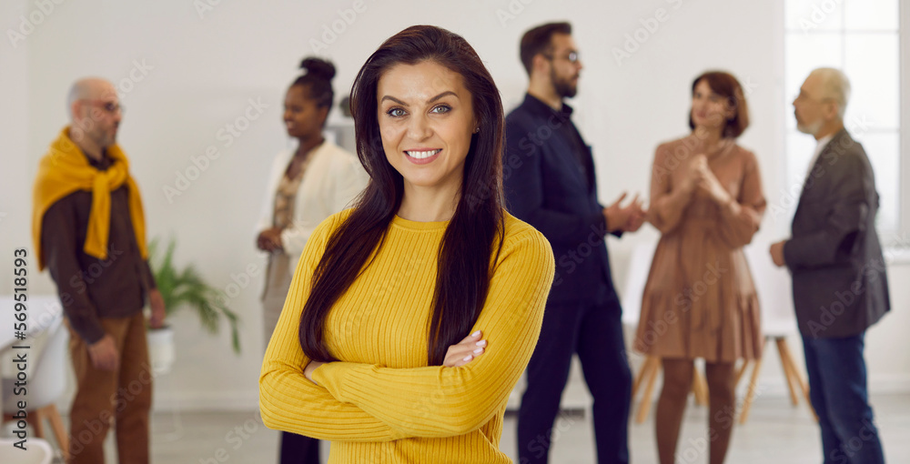 Obraz premium Portrait of successful female employee and business professional at work. Happy beautiful young brunette woman in yellow jumper standing in office with her arms crossed, looking at camera and smiling