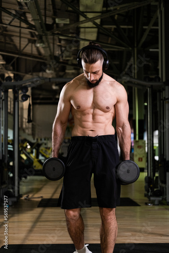 A brutal muscular man with a beard and a perfect body wearing headphones does exercises with dumbbells in the gym. Fitness  beauty