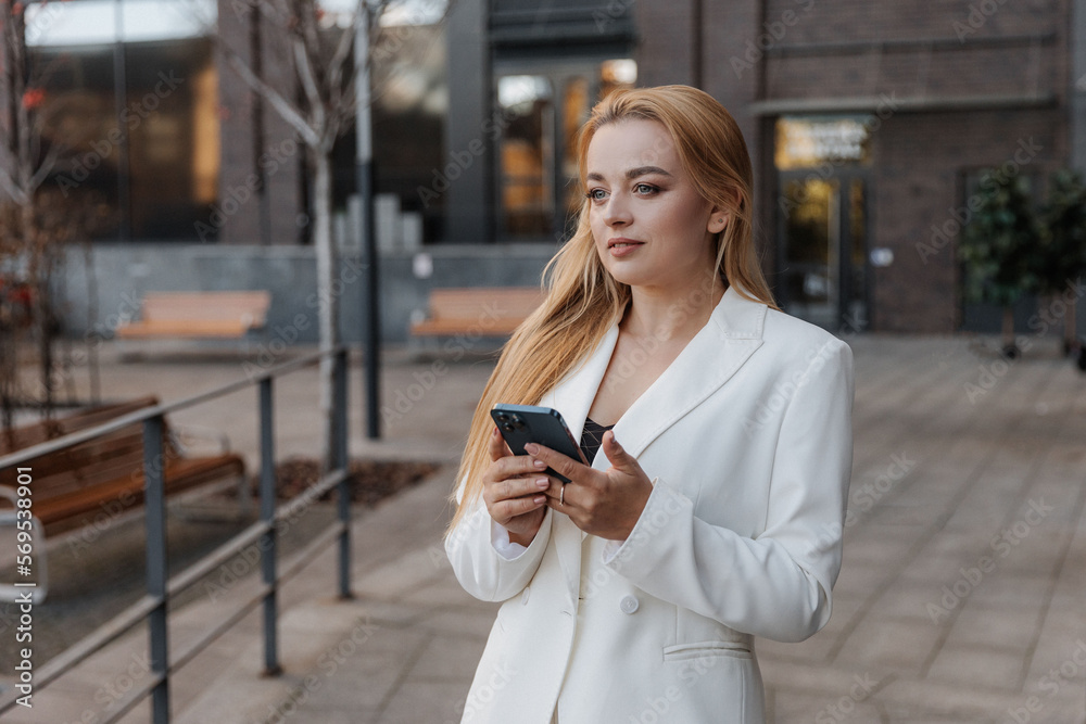 Blond stylish woman at oversized white jacket using phone on the modern city background