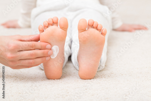 Young adult mother hand applying white moisturizing cream on child foot on on light carpet at home room. Care about children soft legs skin. Closeup. Front view.