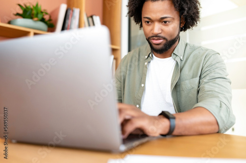 Handsome young businessman is using laptop indoors. African-American curly guy office employee is typing on the keyboard, replying to emails, male freelancer in casual wear working remotely