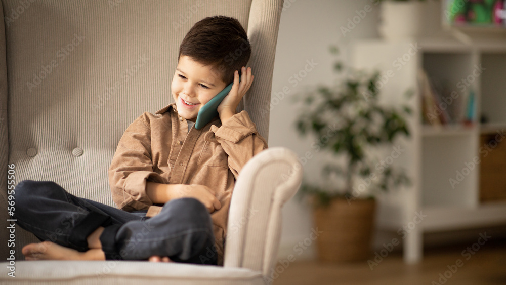 Glad caucasian little kid in casual calls by phone, talk, sits in armchair at spare time in living room interior Glad caucasian little kid in casual calls by phone, talk, sits in armchair at spare time in living room interior
