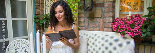 A young smiling woman reading a book and drinking coffee while sitting on her garden patio on a summer day	