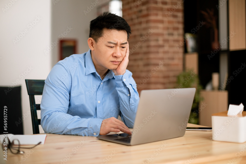 Sad tired middle aged asian man working on laptop computer, sitting at workplace at home office, free space Sad tired middle aged asian man working on laptop computer, sitting at workplace at home office, free space
