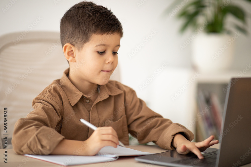 Busy caucasian little child sit at table making notes, watching lesson on computer in school, kindergarten Busy caucasian little child sit at table making notes, watching lesson on computer in school, kindergarten
