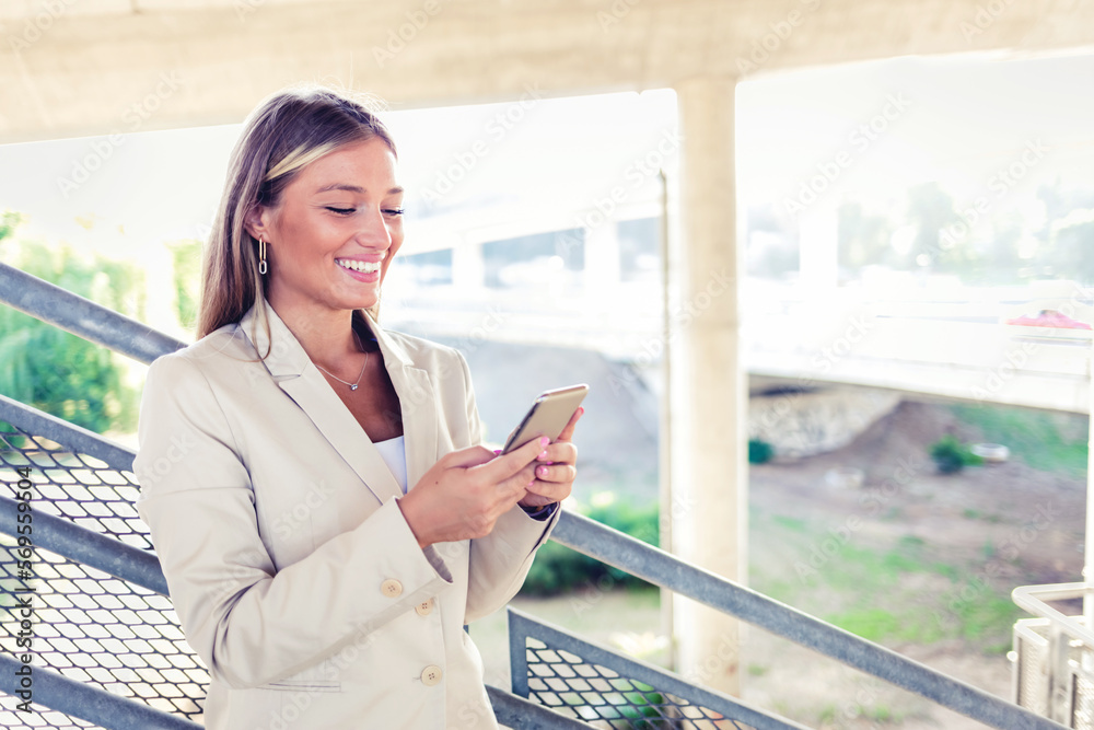 Fototapeta premium Fashionable woman with smart phone. Young businesswoman walking outdoors and using cellphone. Female business professional walking outside and texting from her mobile phone.