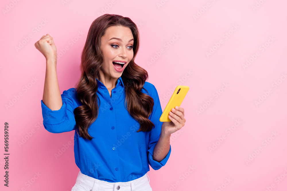 Portrait of adorable nice cute positive girl wavy hairstyle wear blue shirt clenching fist look at phone isolated on pink color background Portrait of adorable nice cute positive girl wavy hairstyle wear blue shirt clenching fist look at phone isolated on pink color background