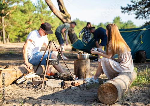 A guy and a young girl sit on a log in nature and talk against the backdrop of scum and put up a tourist tent. The concept of summer recreation in nature on the sandy bank of the river, picnic