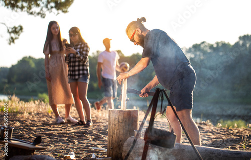 A model caucasian man with a beard and black glasses is chopping wood for a fire at a picnic in the summer. Vacation with friends in nature. Bushcrafting.