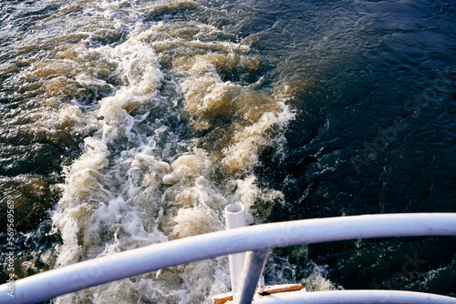 Foamy wake trail on river surface viewed from the deck of a moving boat. Dynamic water motion with visible railing in foreground. Concept of travel, journey, tourism and outdoor adventure.