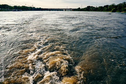 Turbulent river water surface with foam wake trail created by a moving boat. Natural landscape with trees along the shoreline. Dynamic outdoor background representing travel, transportation and nature