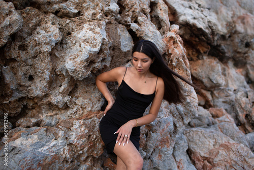 Beautiful brunette in a black dress posing near the rocks and the sea. Attractive brunette woman in a little black dress on the sea coast.