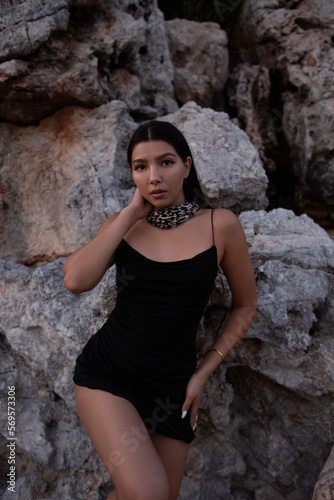 Beautiful brunette in a black dress posing near the rocks and the sea. Attractive brunette woman in a little black dress on the sea coast. © olgamaer
