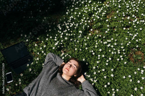 Spring allergies. A beautiful young woman sneezing amidst a blooming flowers, symbolizing spring allergies. Young female laying in green grass next to the laptop. Spring fatigue sleep and calm nap