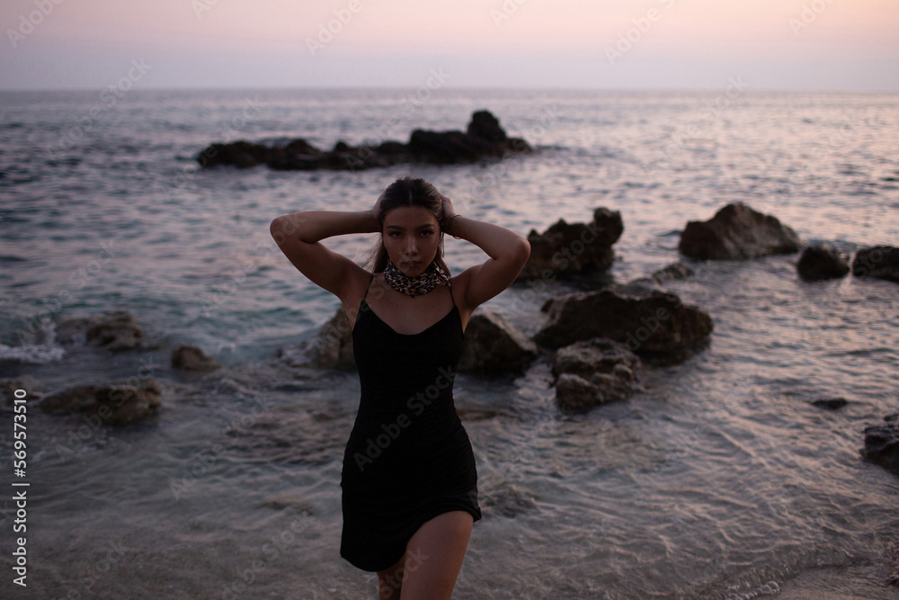 A young girl in a short black dress on the seashore at dusk. Beautiful woman posing in the evening on a beautiful beach after sunset