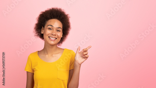 Smiling pretty young african american curly female in yellow t-shirt point finger at copy space