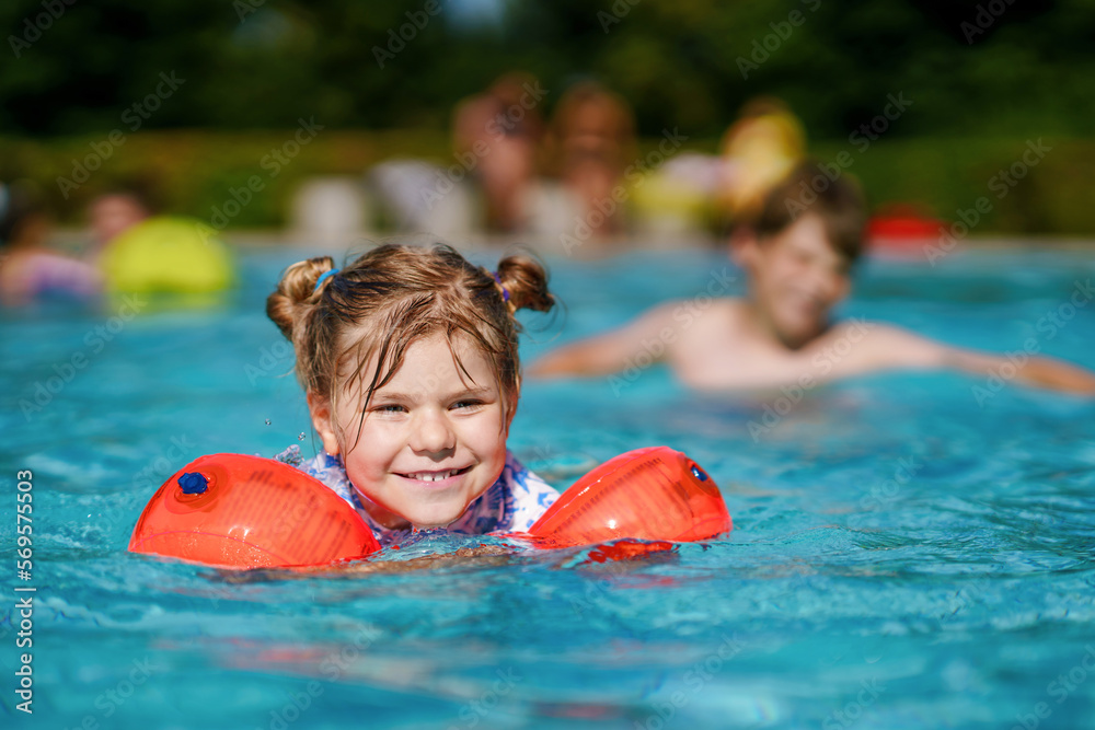 Little preschool girl with protective swimmies playing in outdoor swimming  pool by sunset. Child learning to swim in outdoor pool, splashing with  water, laughing and having fun. Family vacations. Stock Photo