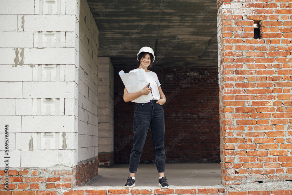Stylish happy woman architect checking blueprints in building house at construction site. Young female engineer or construction worker in hardhat with plans of new modern house