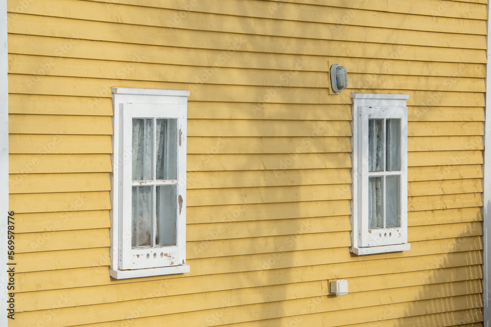 A bright yellow horizontal cape cod horizontal siding wood exterior wall of a cottage with two vintage glass double hung windows. There's a shadow of a tree cast on the exterior wall of the house.