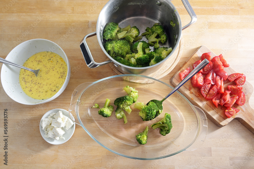 Broccoli in a pot, sliced tomatoes and onions, feta cheese and an egg mixture are filled into a glass baking dish, cooking preparation for a vegetable casserole, high angle view Broccoli in a pot, sliced tomatoes and onions, feta cheese and an egg mixture are filled into a glass baking dish, cooking preparation for a vegetable casserole, high angle view
