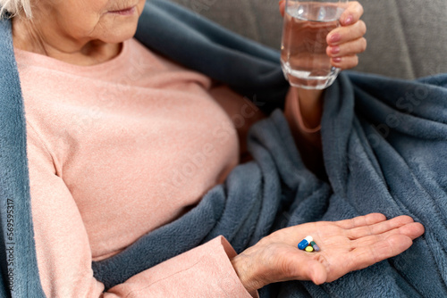 Cropped view of the senior woman holding glass of water and looking on pills while laying at the sofa at home. Age, medicine, healthcare and people concept