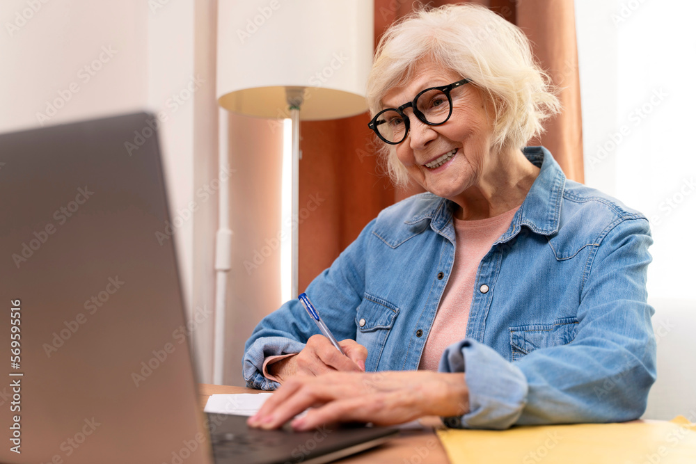 Happy senior woman writing notes while working at home with laptop. Smiling cool old woman wearing eyeglasses teaching students online. Beautiful stylish elderly lady browsing site on computer Happy senior woman writing notes while working at home with laptop. Smiling cool old woman wearing eyeglasses teaching students online. Beautiful stylish elderly lady browsing site on computer