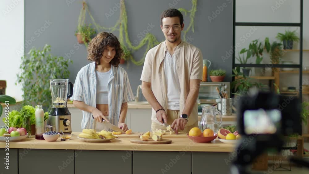 Selective focus shot of young couple cutting fresh fruit and speaking while filming healthy vegan food vlog with digital camera in kitchen at home Selective focus shot of young couple cutting fresh fruit and speaking while filming healthy vegan food vlog with digital camera in kitchen at home