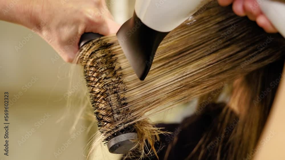 Female hairstylist works with long blonde highlighted lady hair using round brush and dryer in beauty salon closeup slow motion
