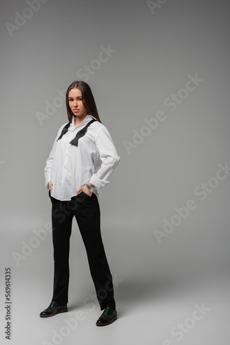 full length of confident woman in shirt with tie and black pants standing with hands in pockets on grey, gender equality concept.