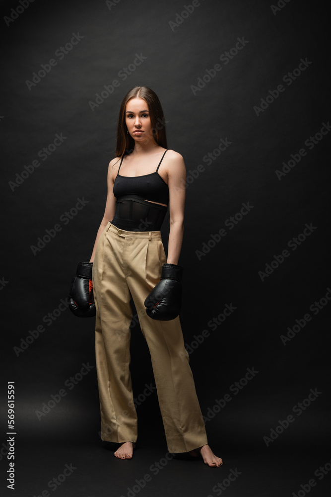 full length of strong woman in top and beige pants posing in boxing gloves on black background. full length of strong woman in top and beige pants posing in boxing gloves on black background.