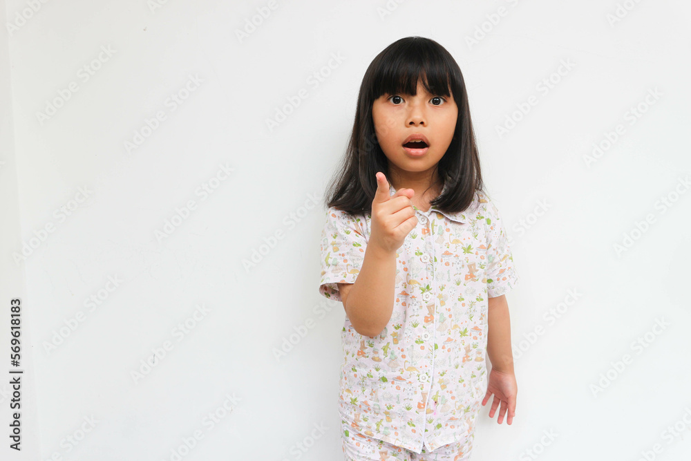 Surprised little girl wearing pajama looking at camera while pointing with hand, isolated on the grey background