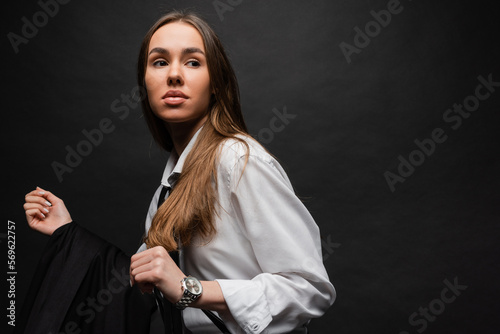 young woman with brunette long hair standing in white shirt and holding blazer on black background.